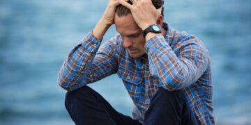 A man in a plaid shirt sits by the water looking distressed, symbolizing stress., tags: gut–brain habits mental - pexel